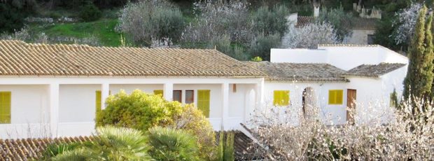 The whitewashed hostal: a long single-storey house with a tiled roof and yellow shutters, almond trees blossoming on the hill behind it.