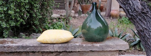 A green glass demijohn and a yellow cushion resting on a worn stone bench, the cactus garden behind.