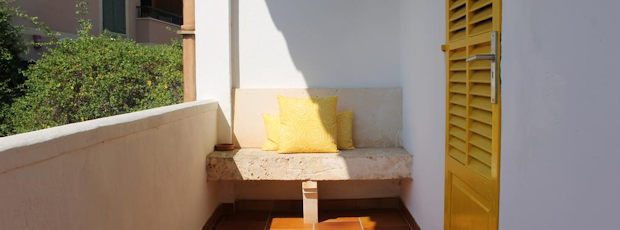 A room's private balcony: a yellow louvred shutter door and yellow cushions on a built-in stone bench in the morning sun.